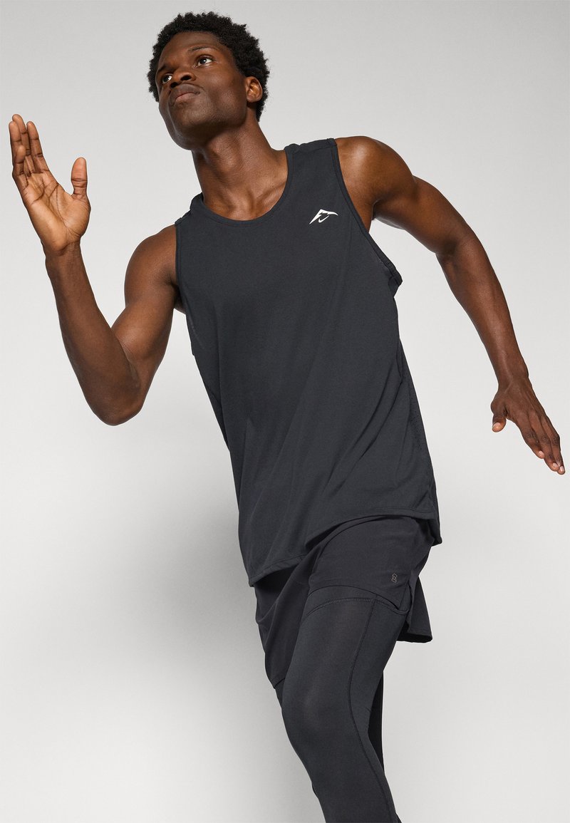 Schwarzes ärmelloses Sporttanktop, aus leichtem Stoff, lockere Passform, mit einem kleinen weißen Logo auf der Brust, kombiniert mit figurbetonenden schwarzen Hosen.