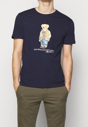 Uomo che indossa una maglietta blu navy con una grafica di un orso in abbigliamento casual e il testo "Polo Bear by Ralph Lauren" sul davanti.