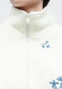 Hvit fleecejakke med høy krage, glidelås foran og blå blomsterbroderier på brystet. Mykt, teksturert materiale.