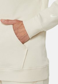 Cream-colored sweatshirt with a side pocket. Features ribbed cuffs and hem. Simple design with a subtle logo accent near the wrist.