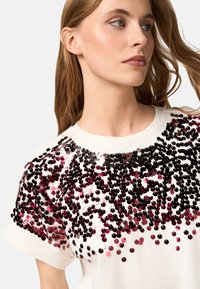 Camiseta blanca con un diseño de lentejuelas en tonos burdeos y rosa, con mangas cortas y escote redondo. Detalle de embellishment texturizado.