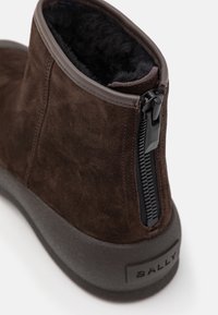 Bottine en daim marron avec doublure en polaire noire, fermeture éclair noire à l'arrière et semelle épaisse en caoutchouc foncé portant la marque "BALLY."