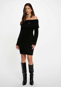 Vestido negro de punto sin mangas con cuello doblado, mangas largas y detalle de botones; combinado con botas de cuero negras hasta la rodilla.