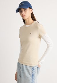 Beige bomull t-skjorte med korte ermer, lagdelt over en hvit langermet skjorte. Lyseblå jeans. Marinblå caps med "Calvin Klein" logo.