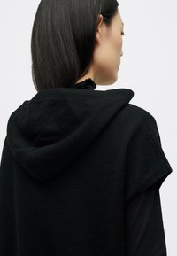 Μαύρο, κουκούλα ένδυμα με κοντά μανίκια, φτιαγμένο από υφαντό ύφασμα. Διακριτές ραφές και χαλαρή γραμμή είναι ορατές από την πίσω όψη.