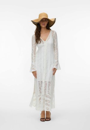 Kvinde iført en lang hvid blondekjole med klokkeærmer og en bredskygget tan solhat, stående foran en ensfarvet hvid baggrund.