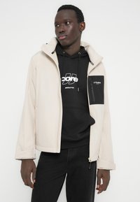 Jeune homme portant une veste beige par-dessus un sweat à capuche noir avec un texte blanc, debout devant un fond blanc uni.