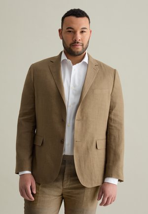 Blazer in lino marrone con rever a punta, due bottoni e taschino sul petto, indossato sopra una camicia bianca, abbinato a pantaloni in lino coordinati.