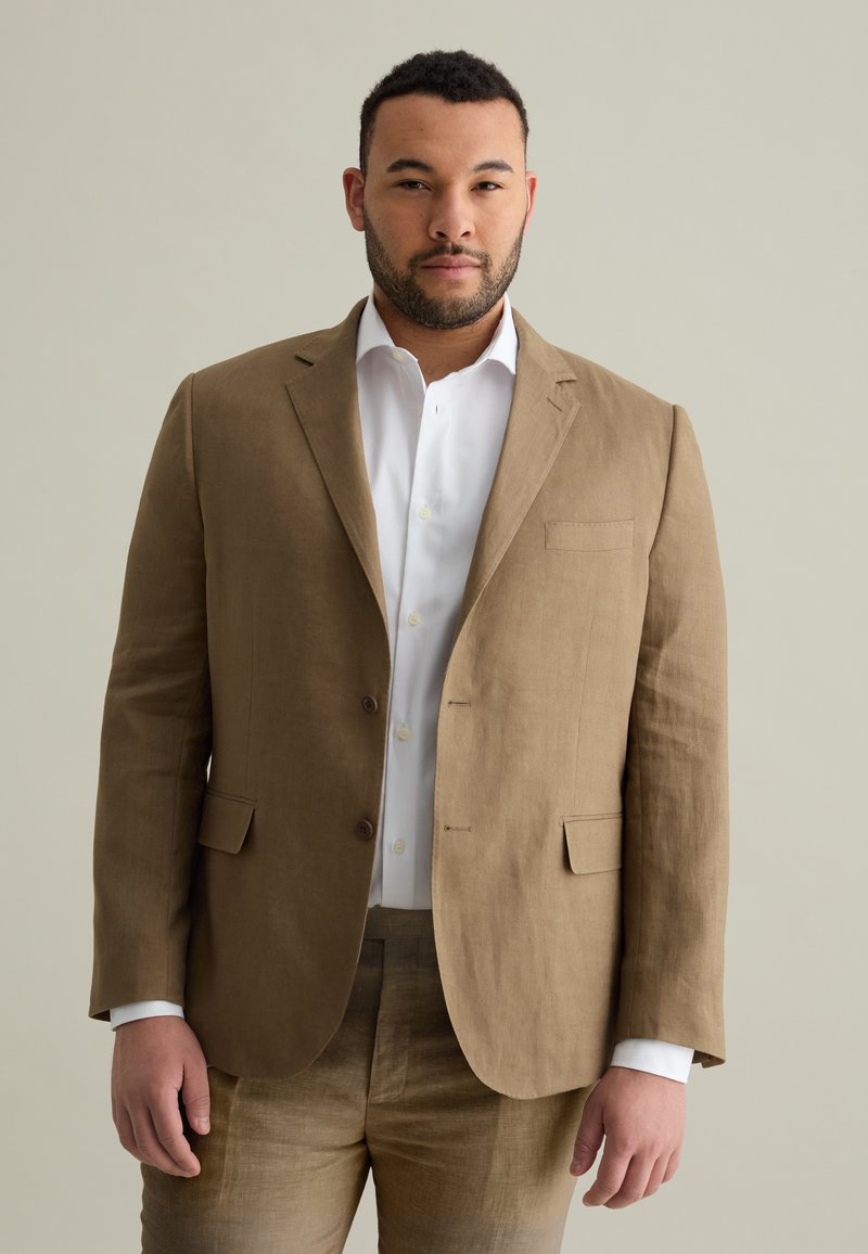 Blazer in lino marrone con rever a punta, due bottoni e taschino sul petto, indossato sopra una camicia bianca, abbinato a pantaloni in lino coordinati.