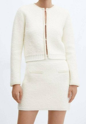 Femme portant une veste courte texturée crème à manches longues et une jupe courte assortie, debout devant un fond uni clair.