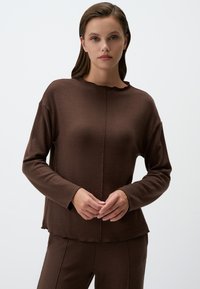 Camicia marrone a maniche lunghe con un colletto leggermente scallopato, tessuto morbido e dettagli cuciti. Visibile abbinata a pantaloni coordinati.