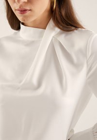 Femme portant un chemisier blanc à col haut avec un détail drapé sur l'épaule droite et des manches longues.