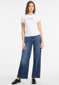 Valkoinen istuva t-paita, jossa on musta "Guess Jeans" -logo, yhdistettynä leveisiin sinisiin farkkuihin. Mustat loafersit täydentävät asua.