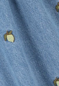 Tissu en denim bleu clair ornée de petits motifs de glands brodés en vert et marron. La texture est douce avec une légère élasticité.