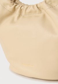 Απαλή μπεζ δερμάτινη τσάντα χειρός με σούρες στο πάνω άκρο και ένθετο χρυσό λογότυπο "COCCINELLE" χαραγμένο κοντά στο κάτω κέντρο.