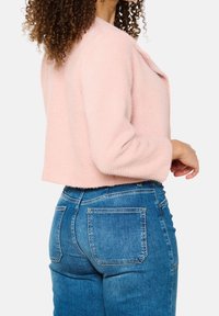 Jasnoróżowy krótki sweter z teksturowanej tkaniny, z luźnymi rękawami i otwartym przodem, zestawiony z niebieskimi, wysokim stanem jeansami.