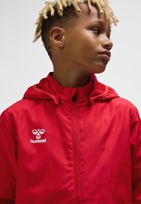 Red, lightweight jacket with a zip closure and a hood. Features a logo on the chest. Smooth texture and minimal design.
