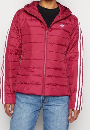 Person iført en vinrød quiltet jakke med hvide striber på ærmerne og lille logo på brystet, kombineret med lyseblå jeans.