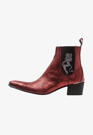 Stivaletto alla caviglia rosso metallizzato con texture, punta appuntita, pannello laterale elastico nero con grafica donna diavolo argento e tacco basso a blocchetto.