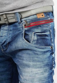 Jeans en denim bleu avec un délavage, poche visible et détails de fermeture éclair rouges. Inclus une ceinture grise avec une étiquette et des accents de boutons en métal.