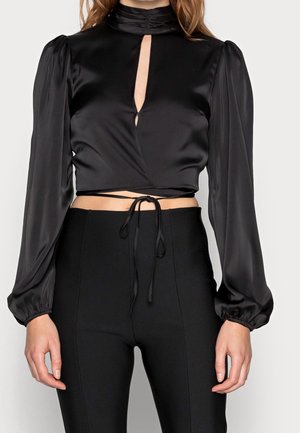 Mujer con un top corto de manga larga de satén negro con escote en forma de ojo de cerradura y detalle de lazo en la cintura, combinado con pantalones negros de talle alto.