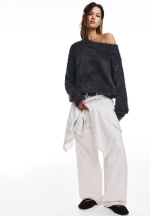 Žena oblečená v tmavém chlupatém svetru s odhalenými rameny, širokých bílých kalhotách, proužkované košili uvázané kolem pasu a tmavých nazouvacích botách pózující na bílém pozadí.