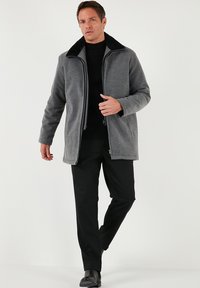 Chaqueta gris de textura suave, con cremallera frontal y cuello de piel sintética negra, combinada con pantalones y zapatos negros, usada por un modelo.