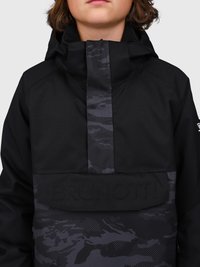 Persona vistiendo una chaqueta con capucha negra con patrones de camuflaje gris oscuro y "BRUNOTTI" estampado en la solapa del bolsillo delantero.