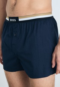 Boxers bleu marine avec une ceinture élastique confortable ornée de rayures dorées, blanches et noires ; fermeture avant à un bouton ; texture de tissu lisse.