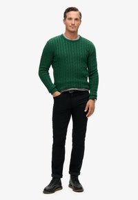 Zielony sweter z splotem w warkocze, okrągły dekolt, ściągacze na rękawach, zestawiony z czarnymi spodniami i czarnymi butami. Mężczyzna stoi z rękami w kieszeniach.