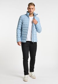 Giacca puffer azzurro chiaro con colletto alto, con un design a righe orizzontali. Indossata sopra una maglietta bianca con jeans neri e sneakers bianche.
