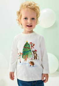 T-shirt blanc à manches longues avec un imprimé festif d'arbre mettant en avant des animaux, des couleurs vives et des détails décoratifs. Porté avec un jean bleu.