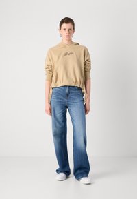 Beige hettegenser med elastisk linning og broderte detaljer, kombinert med blå vide jeans og hvite joggesko. Enkelt design, myk tekstur.