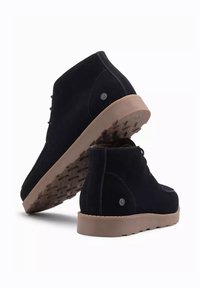 Bottines en suède noir avec une semelle en caoutchouc contrastée, une texture lisse et un design minimaliste, ornées d'un subtil logo sur les côtés.