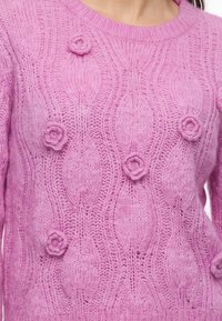 Pull rose tricoté avec des motifs en torsade et de petits détails de fleurs en 3D sur le devant et manches longues.