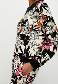 Personne portant un sweat-shirt et un pantalon assortis à imprimé floral noir, blanc, rose et orange, main dans la poche du pantalon, sur fond uni.