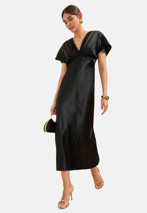 Kvinde i sort satin midi-kjole med blonderkant, guldbøjler, armbånd, hæle og bærende en lille sort håndtaske.