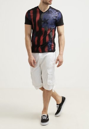 Homme portant un t-shirt noir, rouge et bleu à motifs d'étoiles, un short cargo blanc et des baskets noires, marchant sur un fond clair uni.