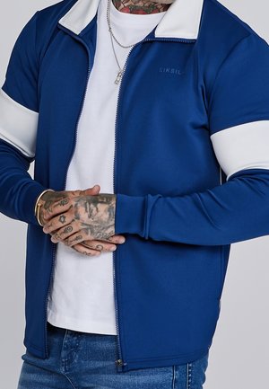 Mand iført blå og hvid lynlåstrøje over hvid skjorte, med tatoverede hænder foldet foran og synlig sølvhalskæde.