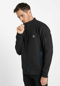 Le sweat-shirt noir présente un col montant, des poches zippées et des fermetures éclair bleues. Le tissu semble doux avec une texture lisse, adapté à une tenue décontractée.