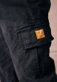 Sorte cargobukser i et tekstureret stof med et fremtrædende orange logopatch og flere multianvendelige lommer. Syninger er synlige på sømmene.