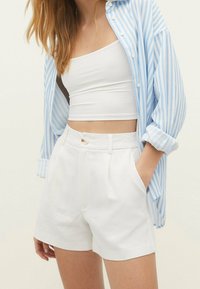 Lichtblauw-wit gestreept shirt met lange mouwen, wit crop top en witte geplooide korte broek. De korte broek heeft een knoopsluiting en zakken.
