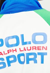 Bílá tkanina s modrým textem "POLO SPORT" a červeným "RALPH LAUREN" uprostřed, se zelenými a modrými barevně blokovanými sekcemi poblíž límce.