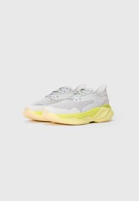 Un paio di sneakers sportive grigie chiare con suole spesse di neon giallo, tomaia in tessuto a rete e chiusura con lacci su uno sfondo bianco.