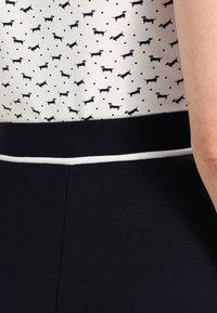 Top a maniche corte realizzato in tessuto bianco con piccoli pois neri e motivi di dachshund ripetuti, complementato da una vita scura con bordo bianco.