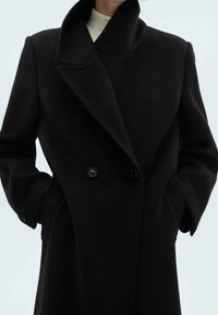 Cappotto di lana nera con colletto alto, design doppiopetto e tasche laterali. Texture liscia con bottoni grandi.