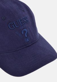 Gorra de béisbol de algodón de color azul marino con logo bordado de "GUESS", detalle de signo de interrogación y agujeros de ventilación a los lados.
