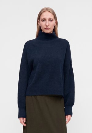 Femme aux cheveux blonds et raides portant un pull à col roulé bleu foncé et une jupe vert olive, faisant face à un arrière-plan uni.