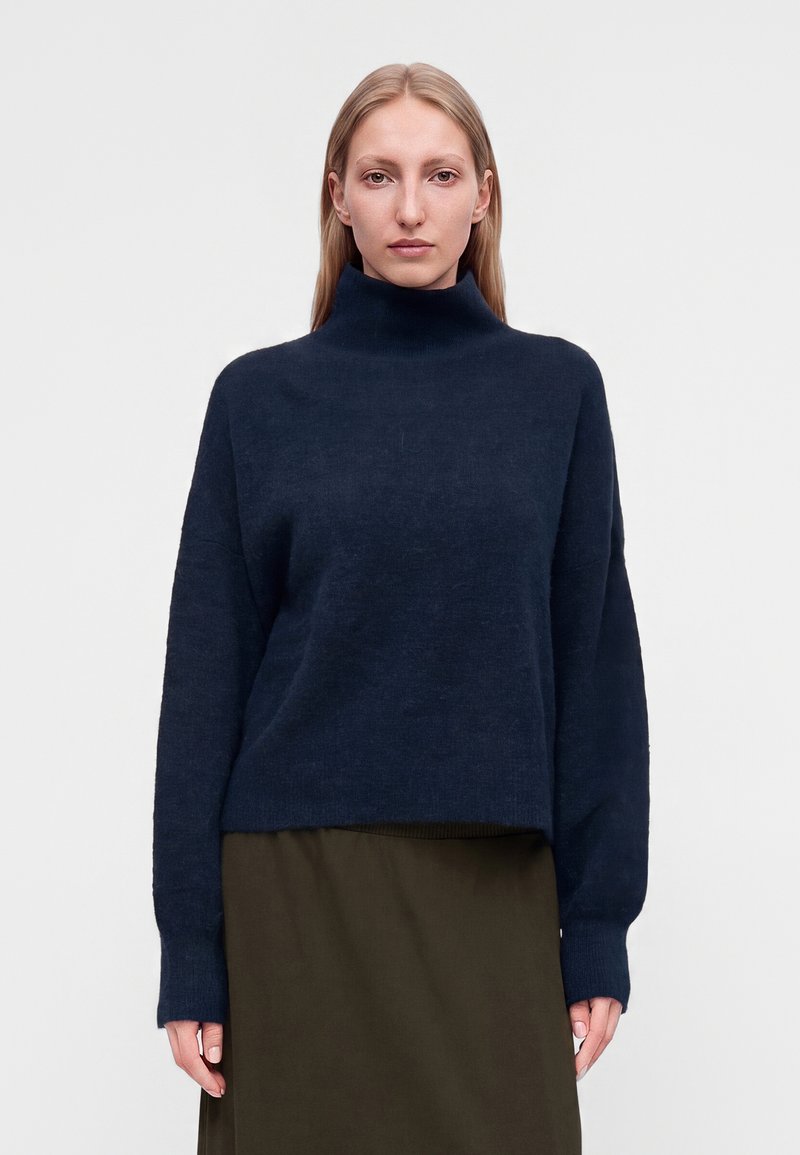 Femme aux cheveux blonds et raides portant un pull à col roulé bleu foncé et une jupe vert olive, faisant face à un arrière-plan uni.