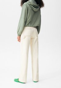 Groene hoodie jas, lichtbeige broek met achterzakken en groene schoenen. Zachte textuur, losse pasvorm, met zichtbare stikseldetails.